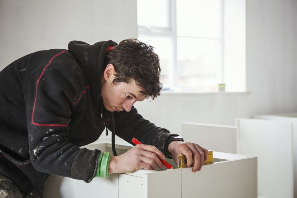 A builder, holding a pencil and ruler, measuring a kitchen unit.